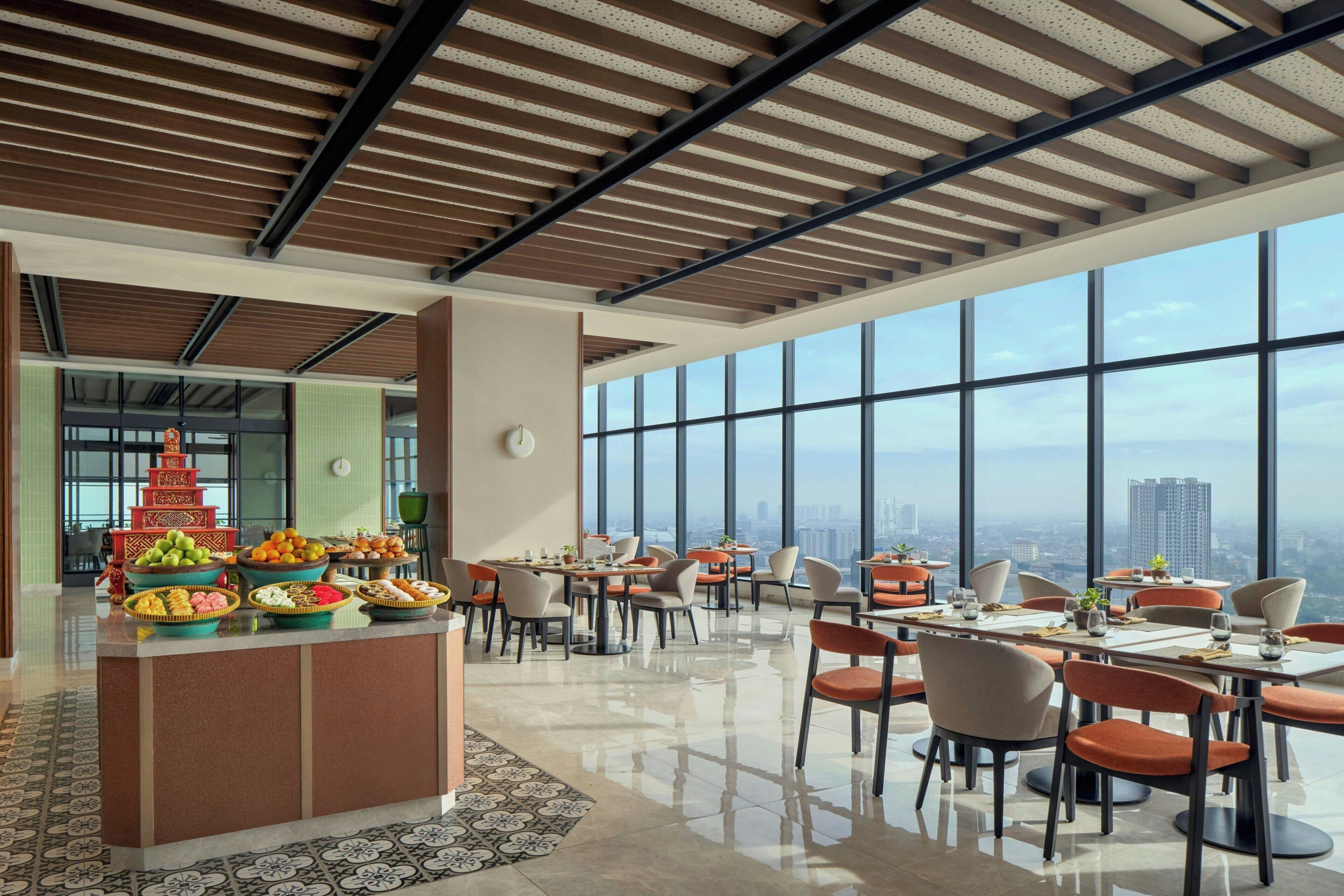A spacious dining area with a high ceiling, large windows offering a city view, and a central buffet with various food items.