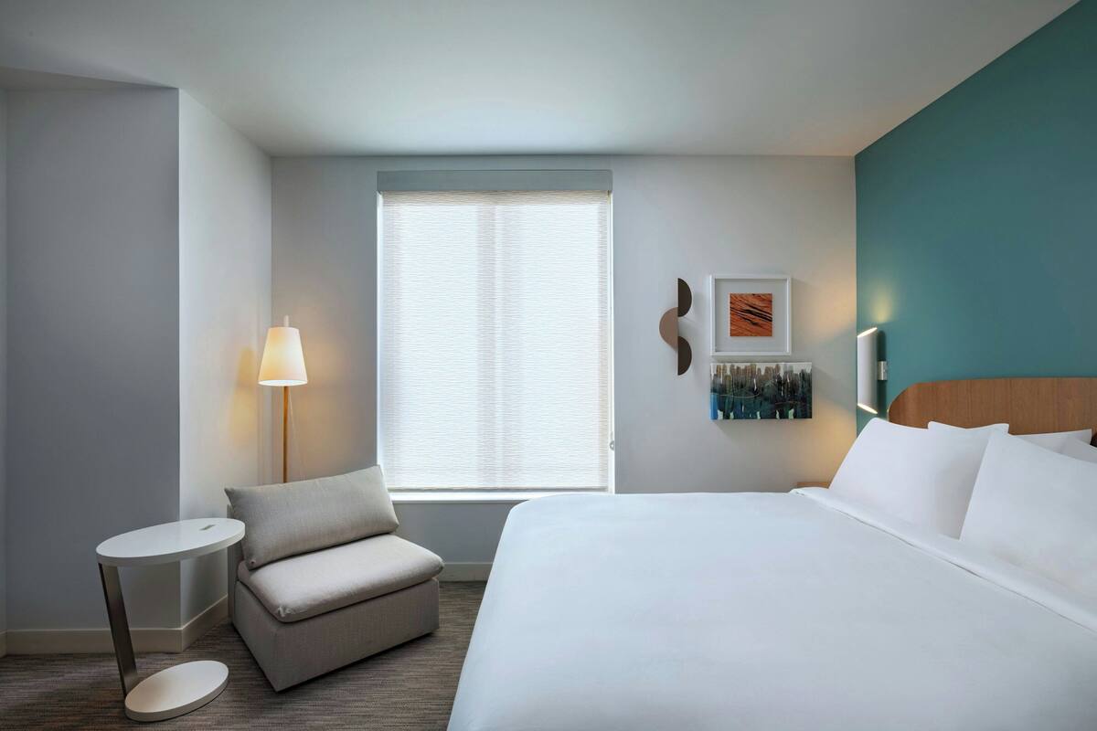 A hotel room with a bed, a small round table, a sofa, a window with blinds, and wall art.