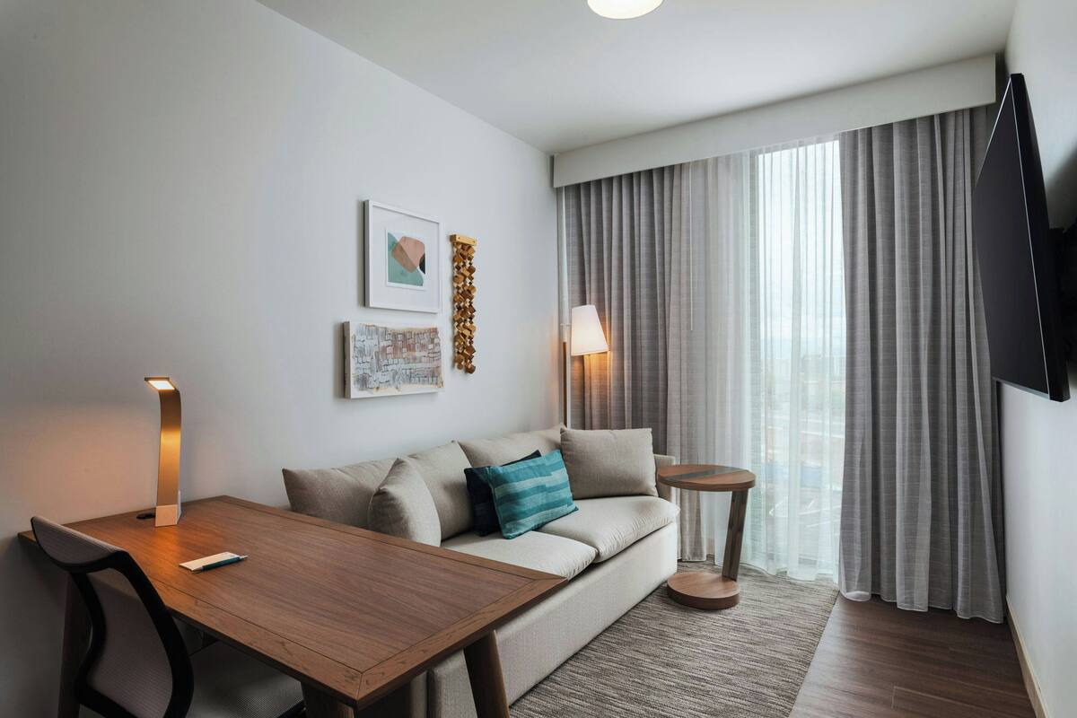 A modern hotel room with a wooden dining table, a sofa with cushions, a small round side table, a lamp, a flat-screen TV, and a large window with curtains.