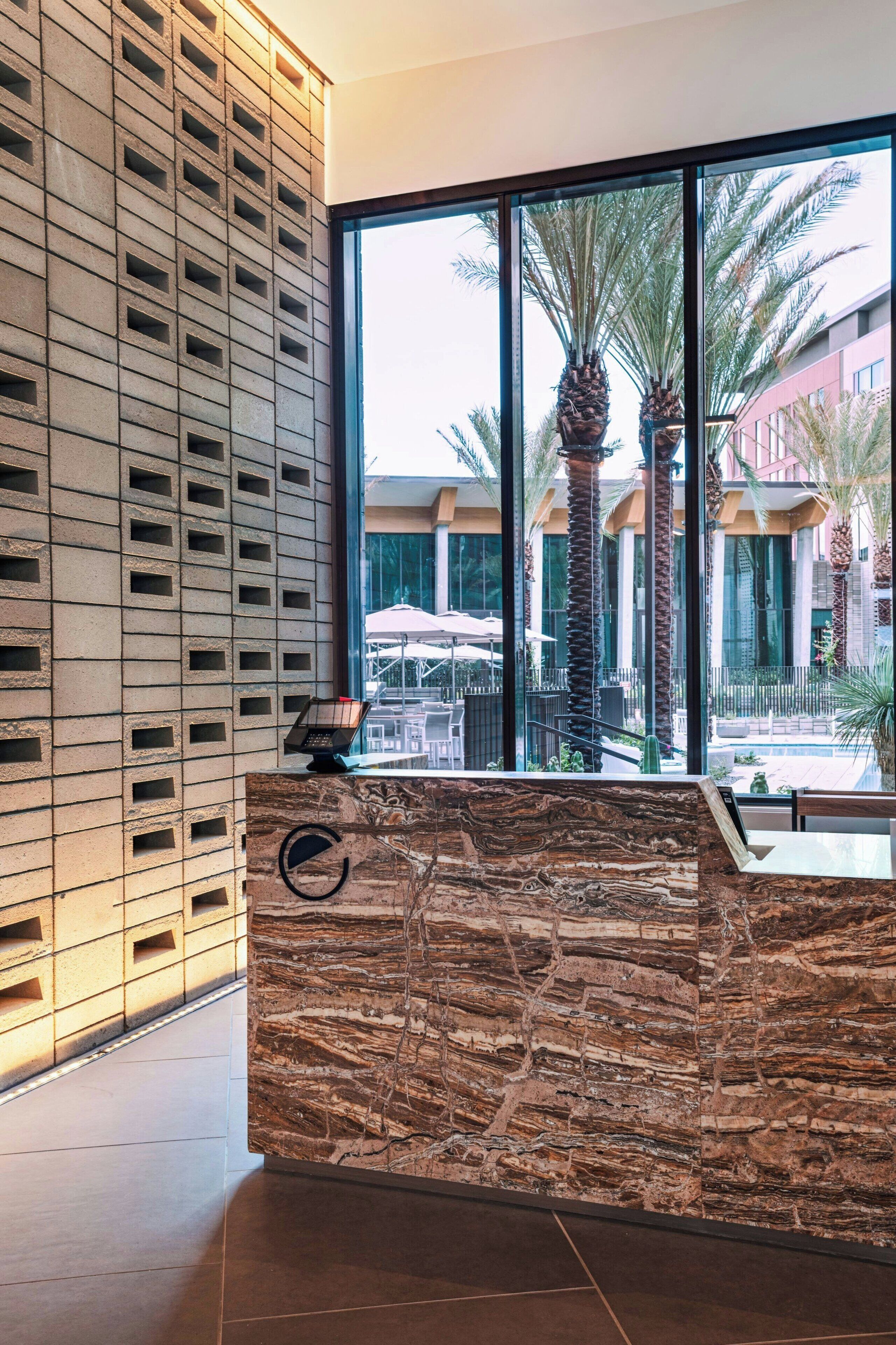 A reception desk with a marble design, a modern wall with a grid pattern, and a view of palm trees and outdoor seating through large windows.