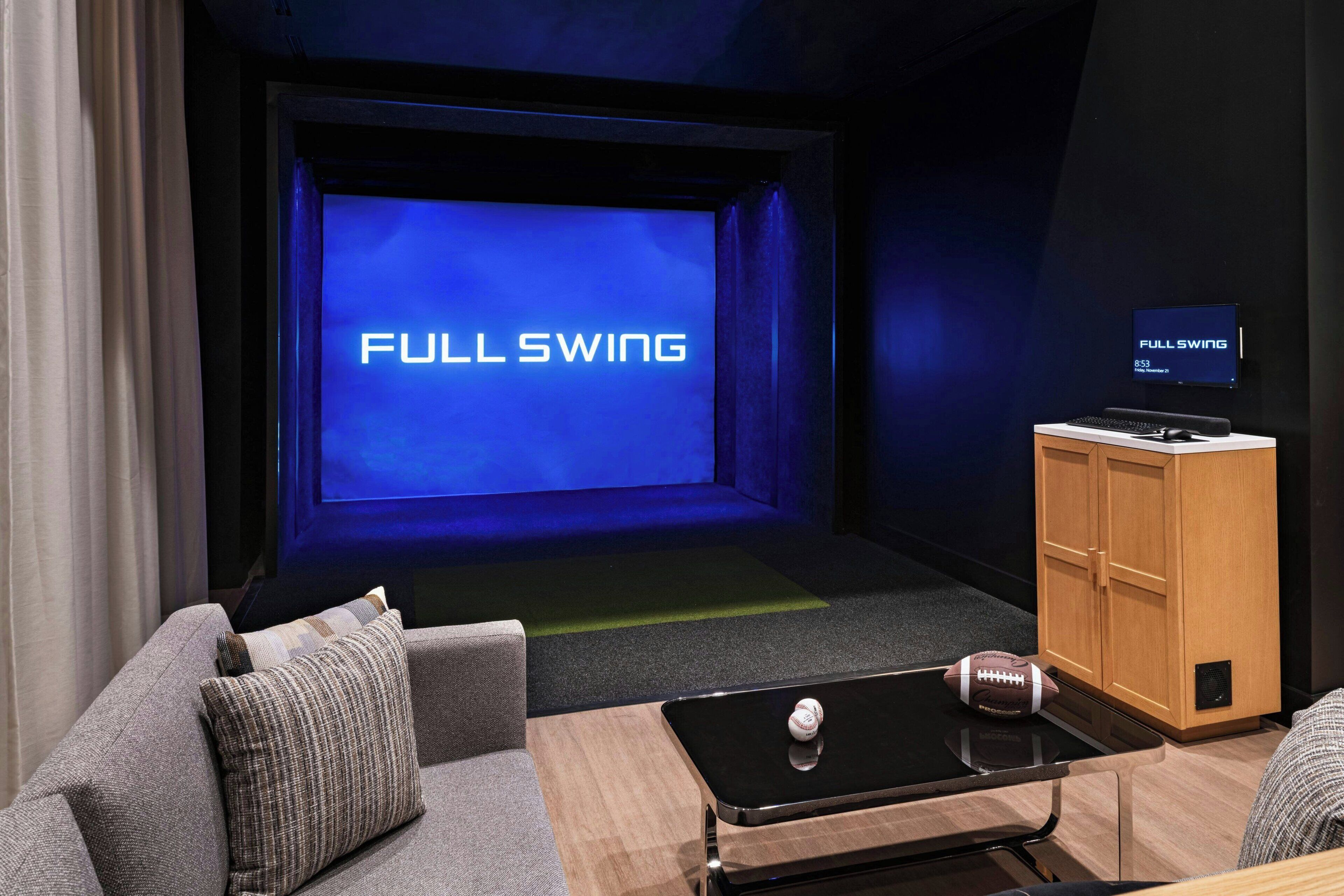 A room with a large screen displaying "FULL SWING," a small wooden cabinet, a couch with a pillow, and a coffee table with a football.