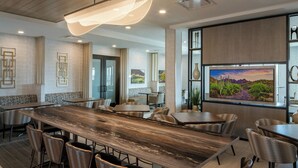 A modern dining area with a long wooden table, multiple chairs, and a large TV screen displaying a desert landscape.