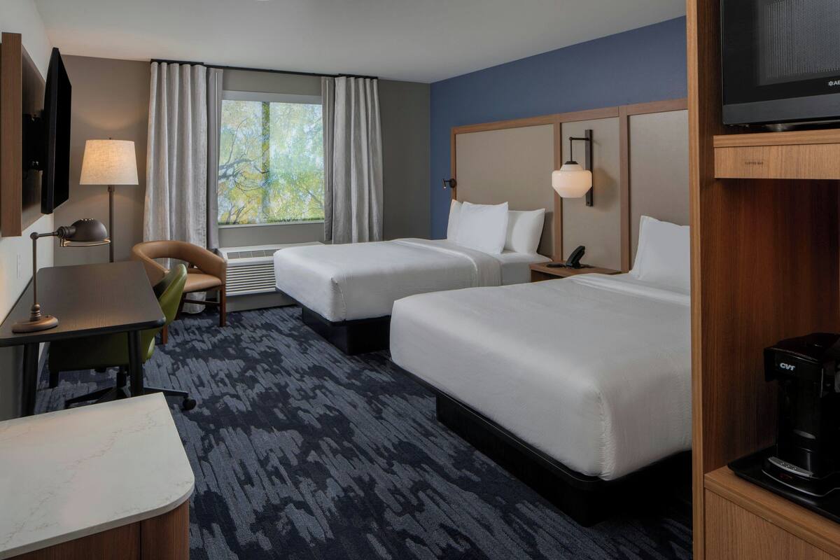 A hotel room with two beds, a desk, a chair, a TV, and a coffee maker.