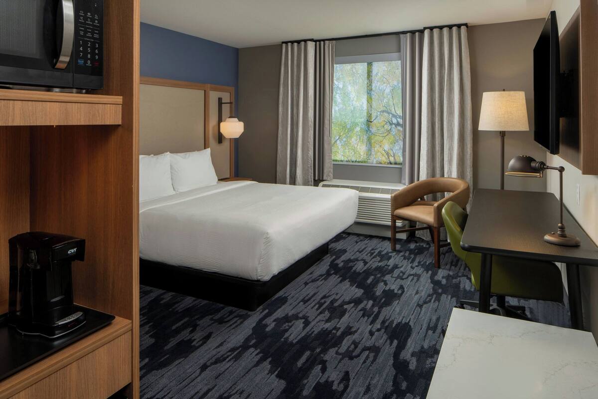 A hotel room with a bed, a desk, a chair, a TV, a coffee maker, and a window with curtains.