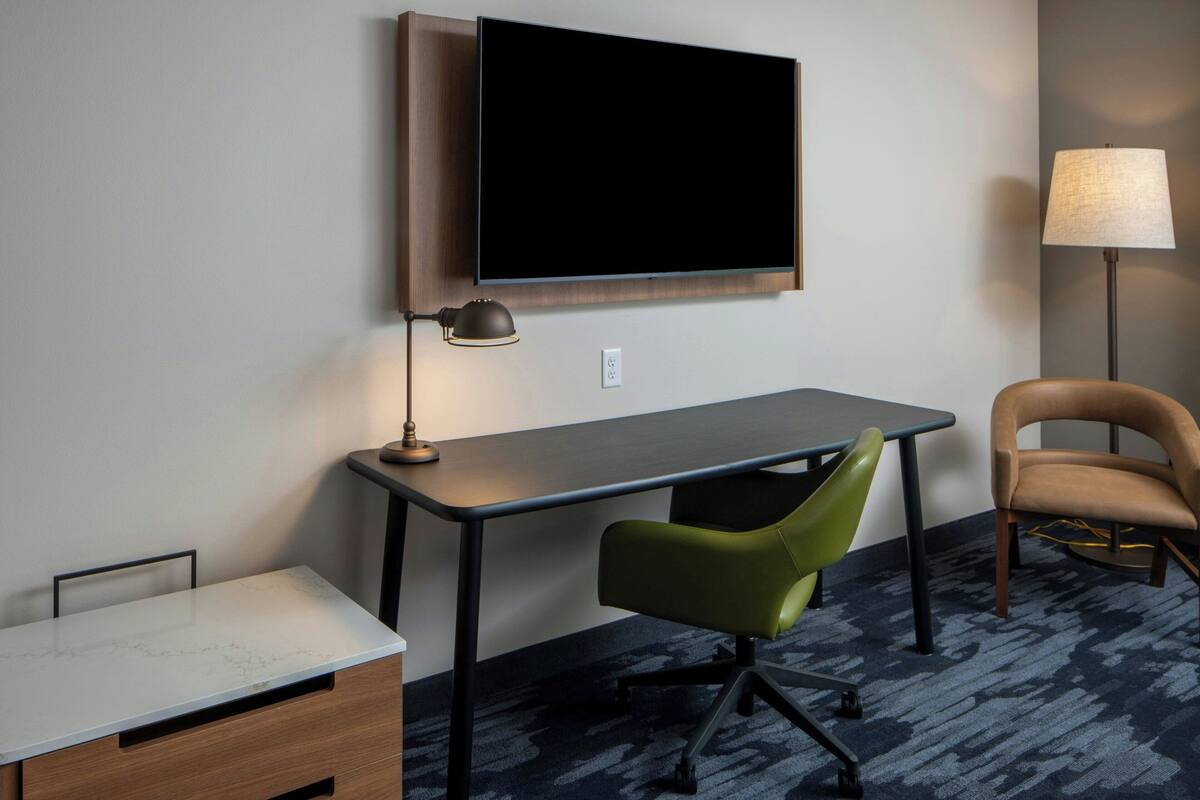 A hotel room with a flat-screen TV mounted on the wall, a desk with a chair, a lamp, and a sofa.