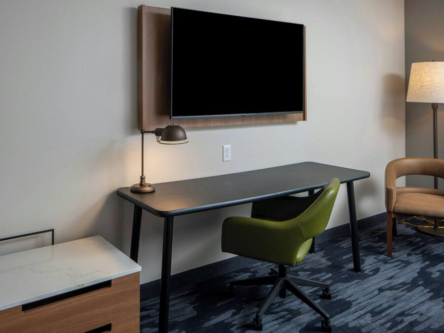 Quarto de hotel com televisão de tela plana na parede, uma escrivaninha com cadeira, luminária e sofá.