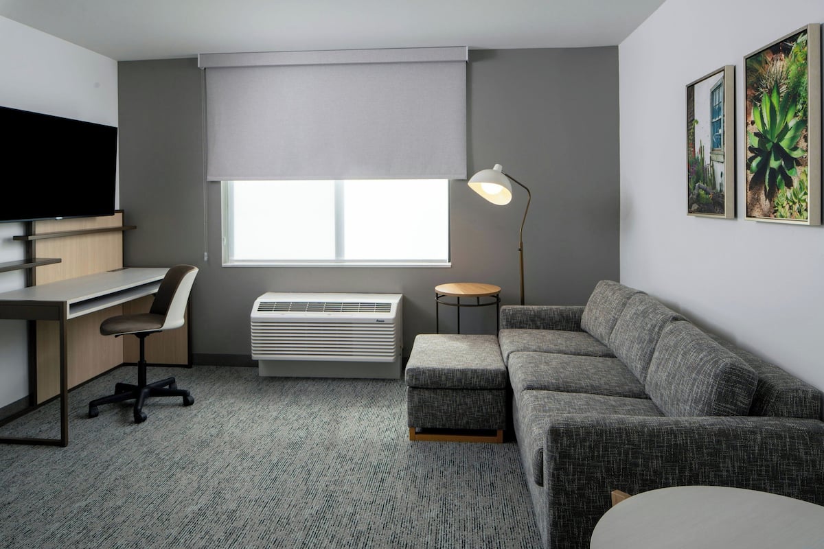 A modern hotel room with a desk, chair, sofa, and a large window with blinds.
