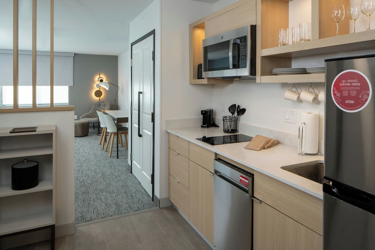 A modern kitchen with a built-in microwave, stainless steel refrigerator, and a coffee maker. There is a dining area with a table and chairs visible through an open door.