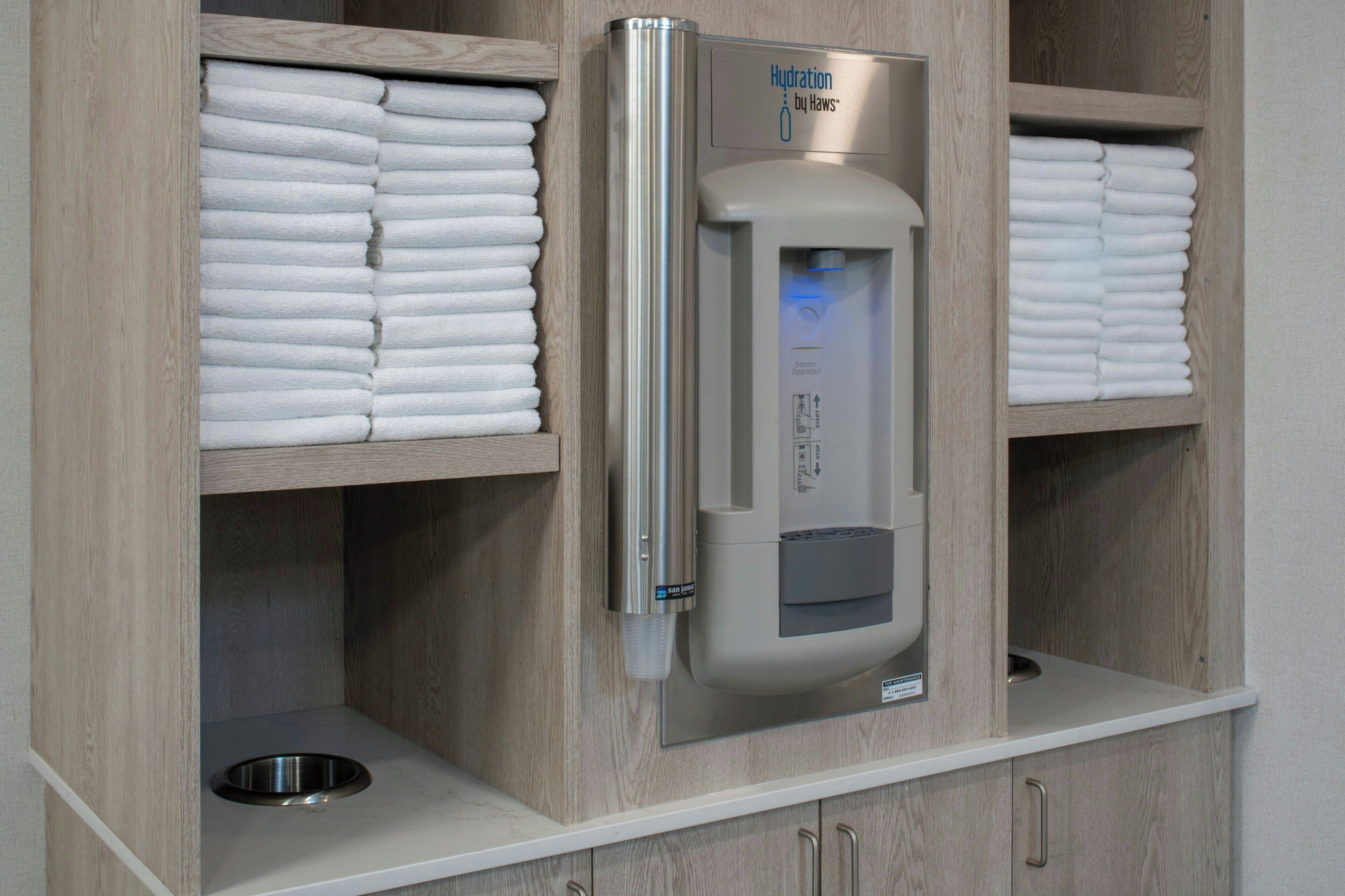 A bathroom with a towel dispenser, shelves with folded towels, and a metal sink.