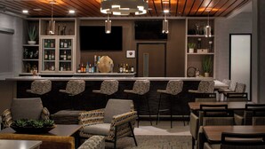 A bar area with a long counter, multiple bar stools, and a variety of bottles and glasses. There are several chairs and tables arranged for seating. - Towneplace Suites By Marriott Surprise Prasada (Surprise)