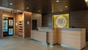 A reception area with a wooden reception desk, a wall-mounted decorative piece, and a refrigerated display case with beverages. - Fairfield By Marriott Inn & Suites Foley (Foley)
