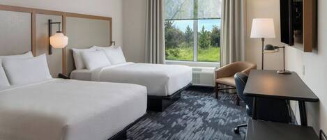 A hotel room with two beds, a desk, a chair, a TV, and a window with a view of greenery.