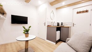 Een compacte woonruimte met een flatscreen televisie aan de muur, een klein rond tafeltje met een potplant, een kitchenette met een magnetron en een kleine koelkast, en een grijze bank.