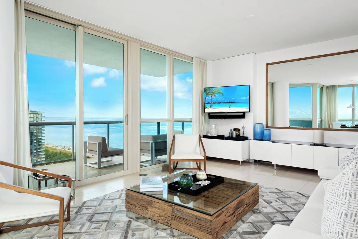 A modern living room with a large glass door leading to a balcony with a view of the ocean. There is a flat-screen TV mounted on the wall, a white sofa, and a wooden coffee table with decorative items.