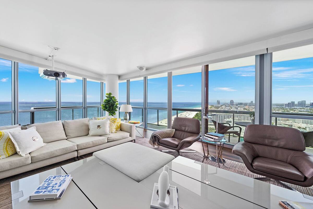 A modern living room with a large sofa, two leather chairs, a coffee table, and a view of the ocean and city skyline.