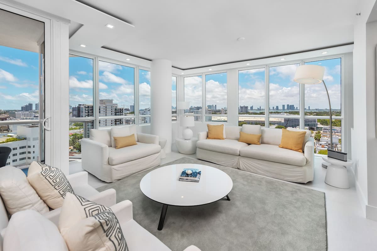 A modern living room with white sofas, a round coffee table, and large windows offering a city view.