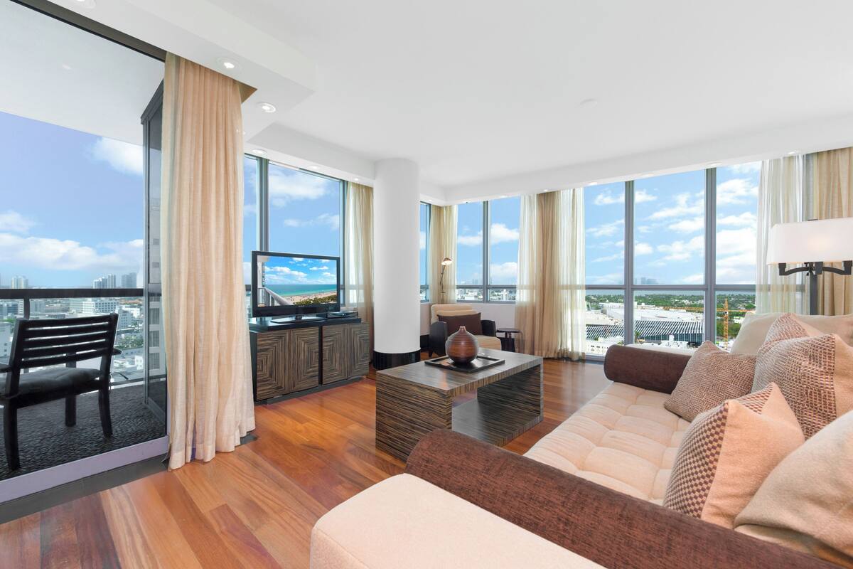 A spacious living room with a large sofa, a coffee table, and a television. There are large windows offering a view of the city.