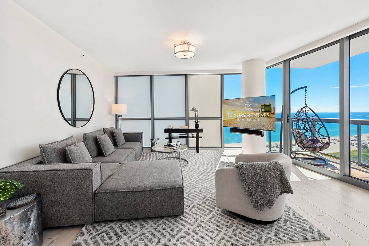 A modern living room with a grey sofa, a small round table, and a TV screen displaying 'Luxury Rentals Miami Beach'.