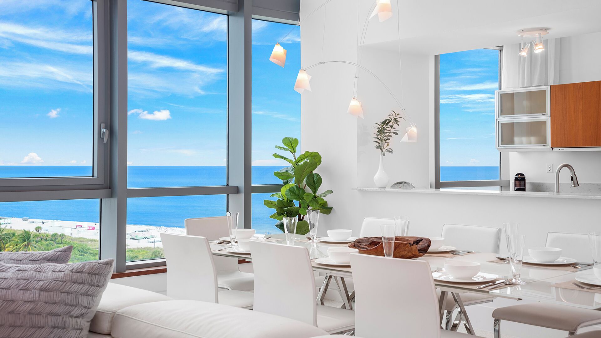 Zona pranzo moderna con vista sul mare, arredi bianchi e illuminazione a sospensione.