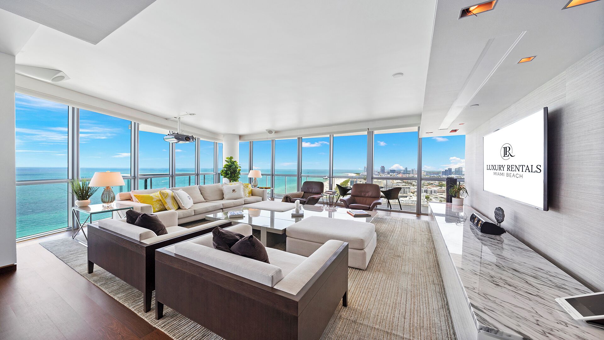 A spacious living room with a large sofa, armchairs, and a coffee table. There is a large window offering a view of the ocean and city skyline. A flat-screen TV is mounted on the wall.