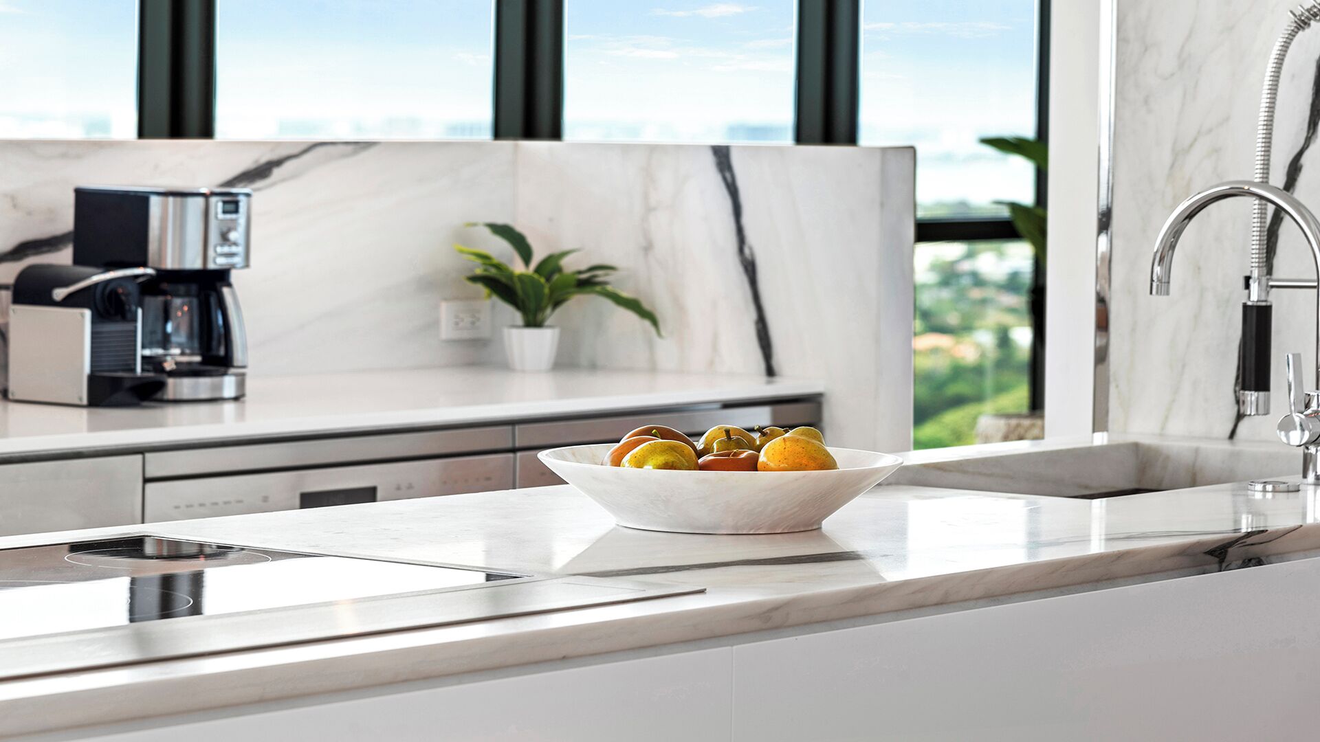 A modern kitchen with a marble countertop, a coffee machine, a plant, and a bowl of fruit.