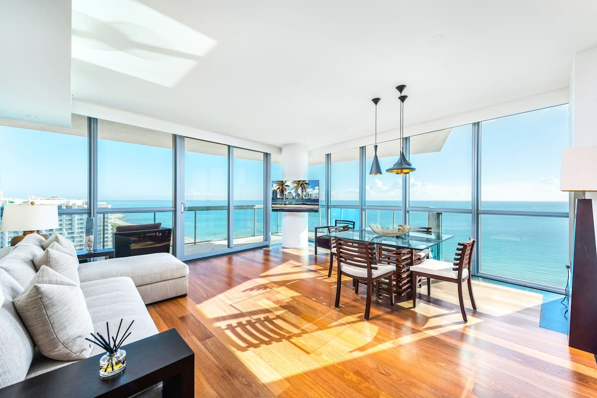 A spacious living room with a large glass table and chairs, a sofa with cushions, and a view of the ocean.
