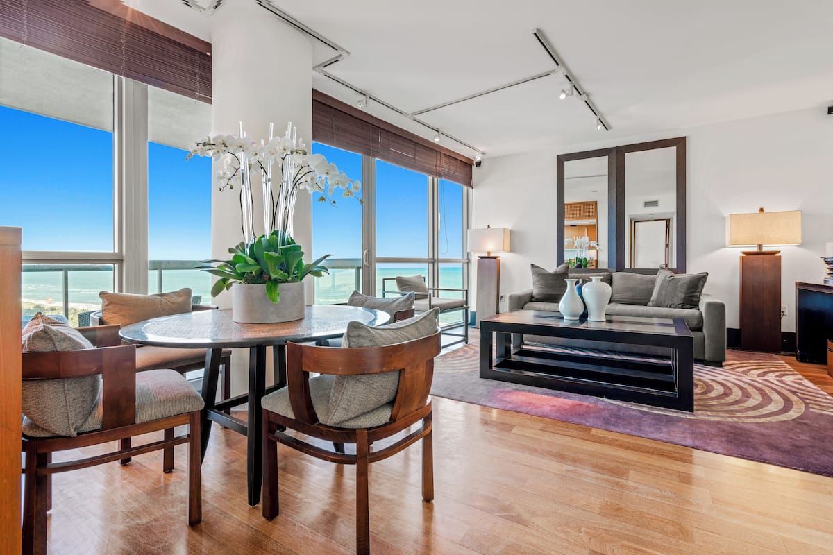 A modern living room with a large window overlooking the ocean, a round dining table with chairs, a sofa, and two lamps.