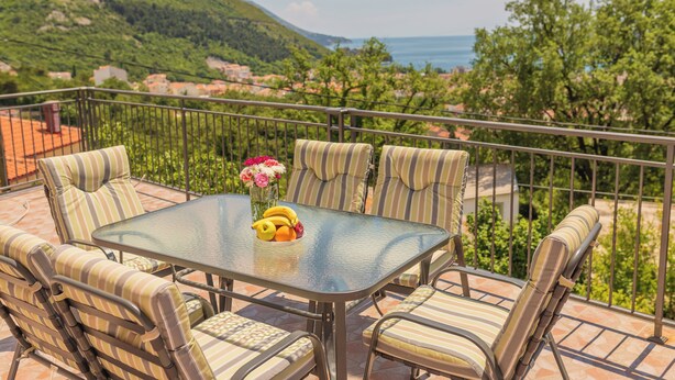 A balcony with a glass table, striped cushioned chairs, and a view of a green hillside and sea. - Villa Sunny Hill (Budva)
