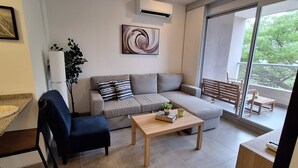 Un salon moderne avec un canapé gris, une table basse en bois, un fauteuil bleu et une grande fenêtre donnant sur un balcon.