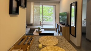 Un salon moderne doté d’un grand téléviseur, d’un coin repas avec une table et des chaises en bois, et offrant une vue sur l’extérieur grâce à de grandes fenêtres.