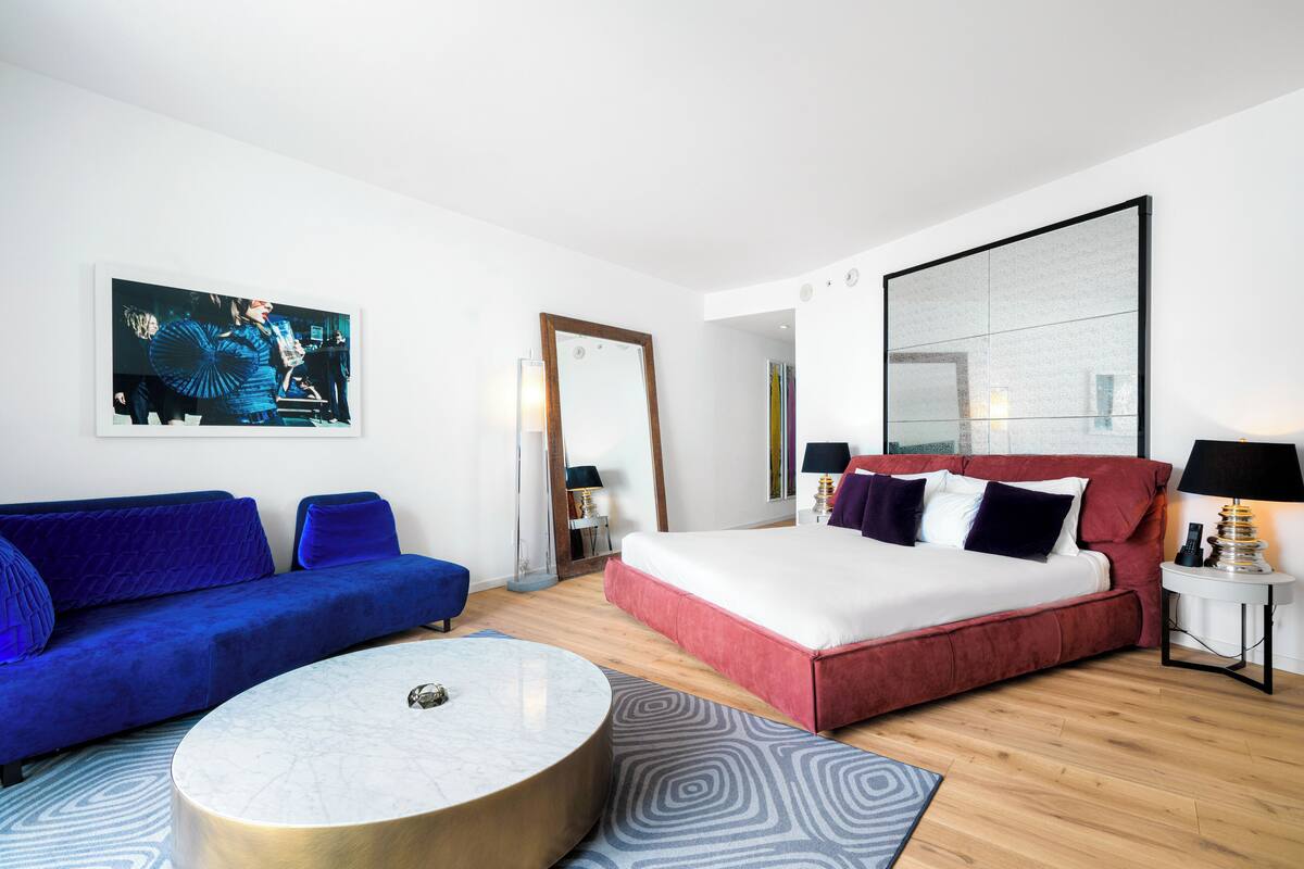 A modern hotel room with a blue sofa, a round coffee table, a red bed, and a large mirror.
