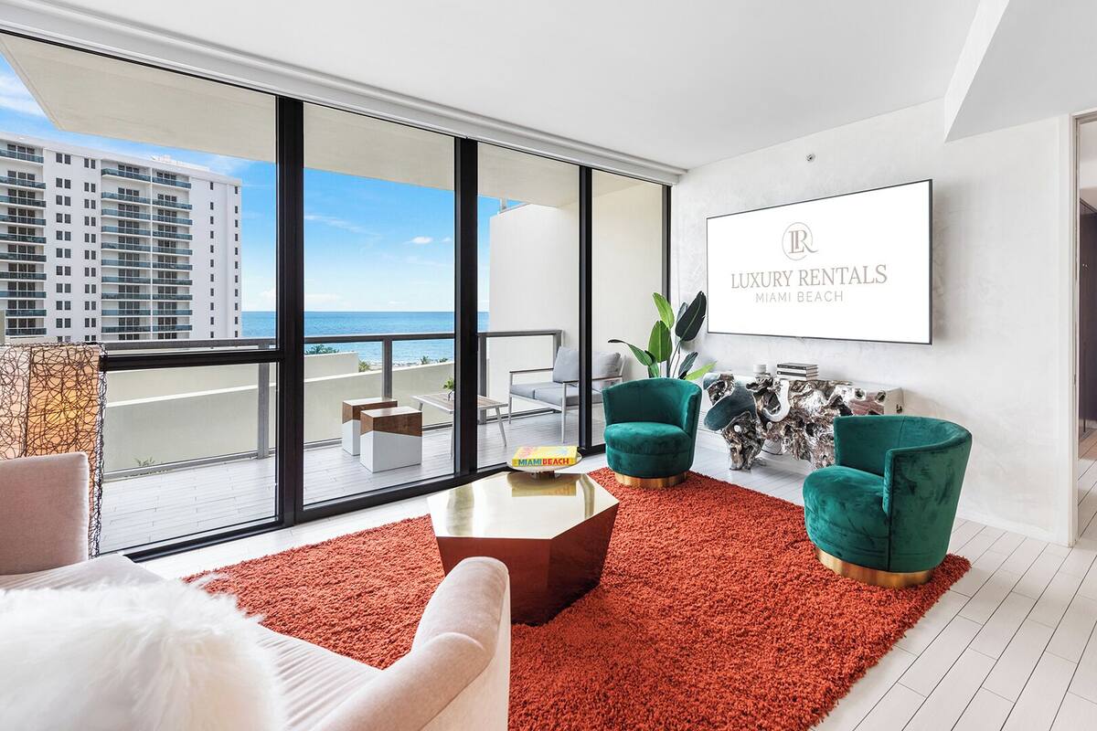 A modern living room with a large window overlooking Miami Beach, featuring two teal armchairs, a white sofa, and a red rug.