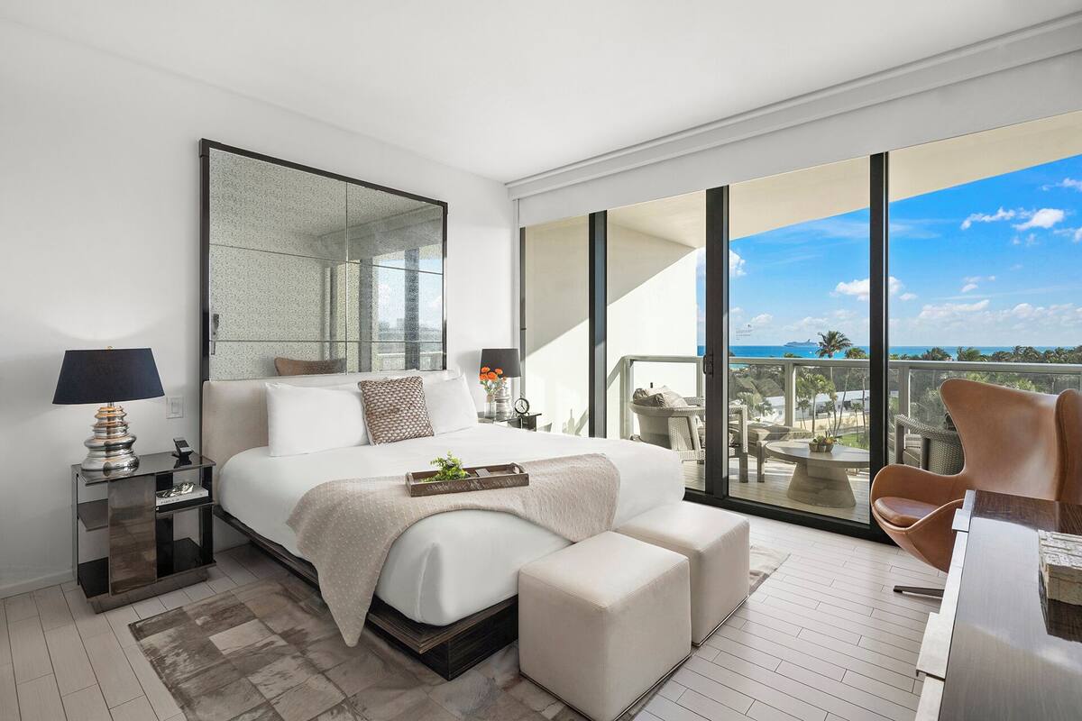 A modern hotel room with a large bed, a sitting area with a chair, a balcony with a view of the ocean, and a large mirror.