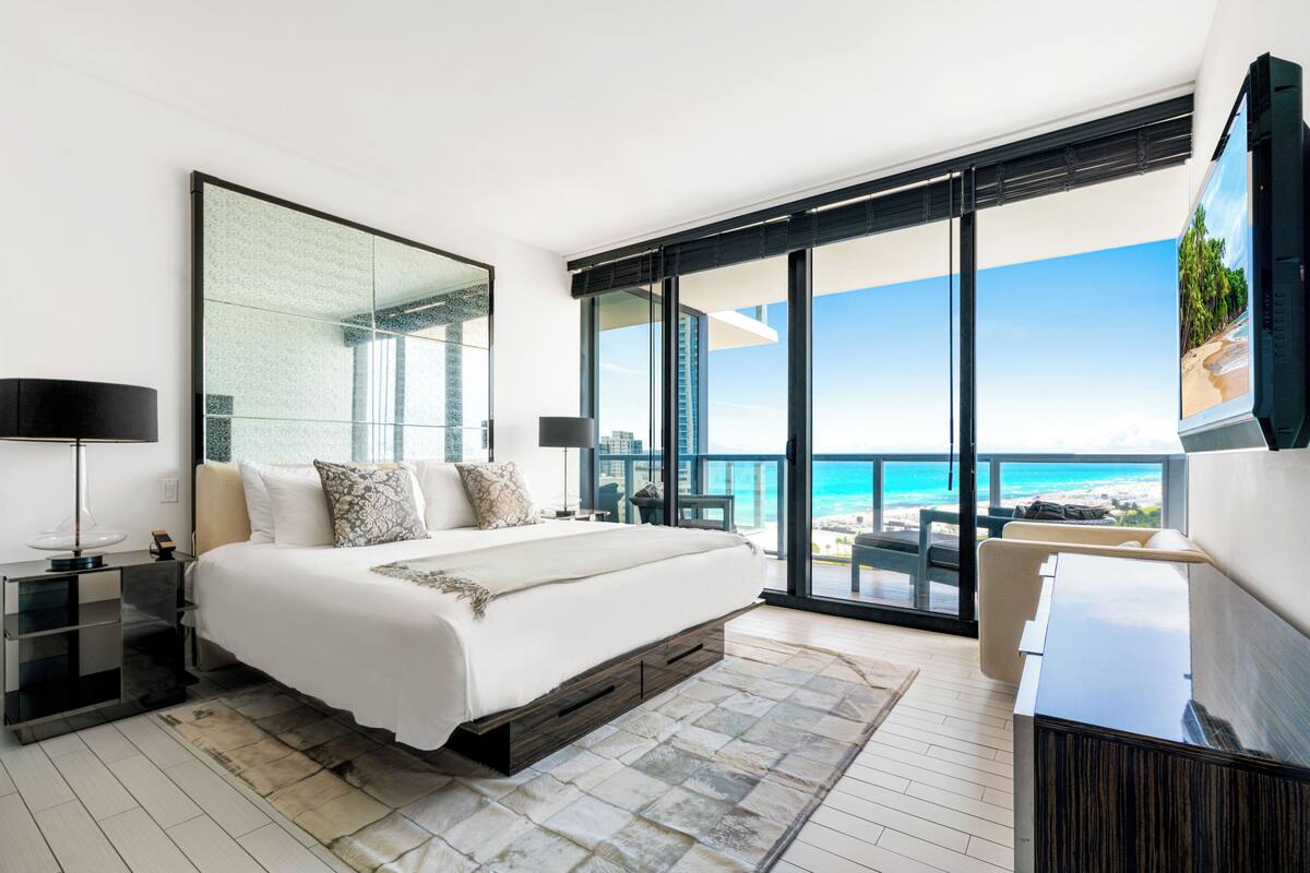 A modern bedroom with a large bed, a TV, and a balcony with a view of the beach and ocean.