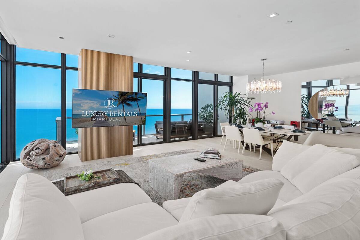 A spacious living room with a large TV, white furniture, and a view of the ocean.