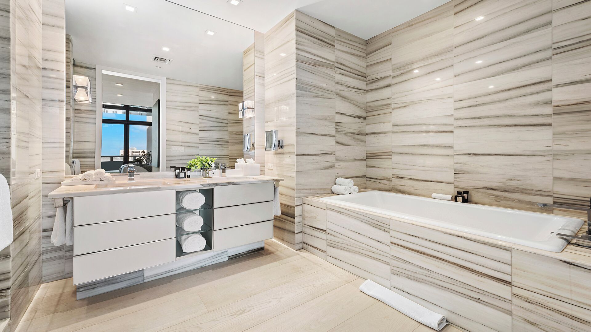 A modern bathroom with a marble wall, a large mirror, a vanity with a sink, and a bathtub.