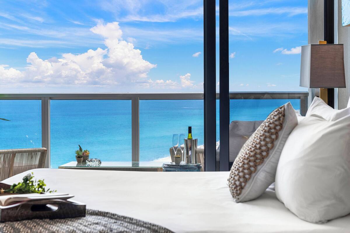A balcony with a view of the ocean, a bed with pillows, a small table with a bottle and glasses, and a wicker chair.