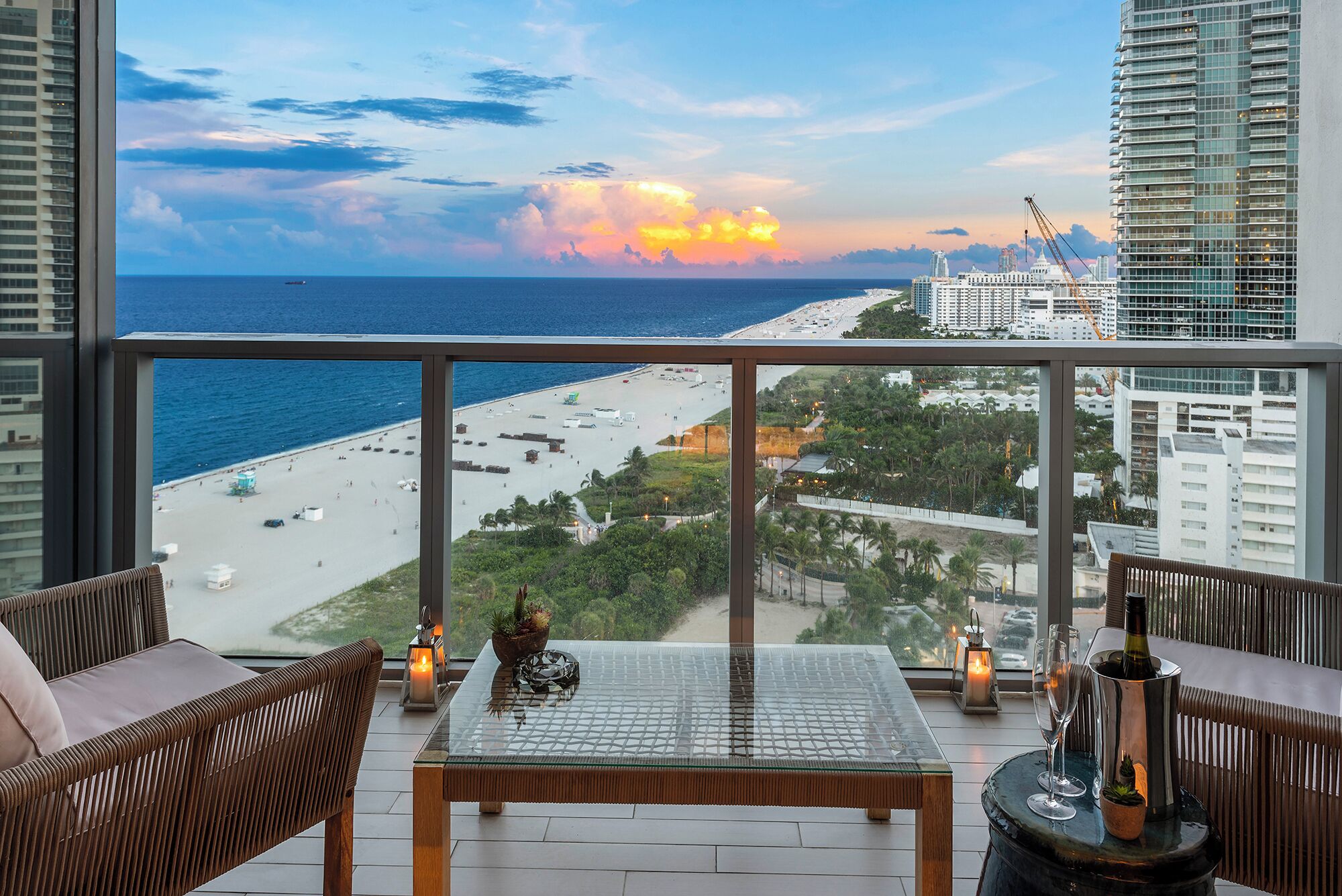 A balcony with a glass table, wicker chairs, and a view of a beach and cityscape during sunset.