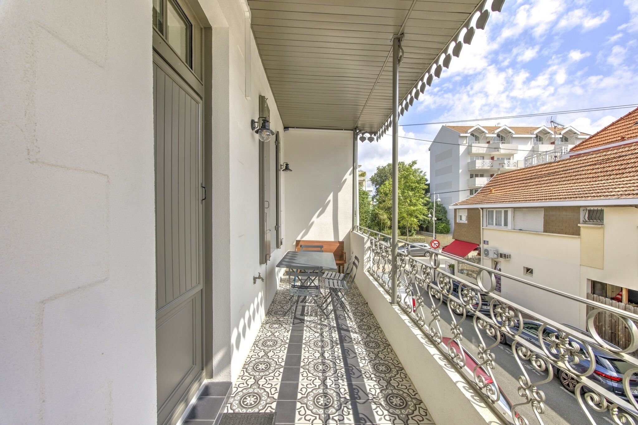 A balcony with patterned tiles, a table and chairs, and a view of a street with parked cars and buildings.