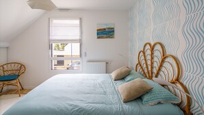 A bedroom with a large bed, a wicker chair, a window with blinds, and a patterned wall. - Ti Armor - Maison à 200m de la plage (Benodet)
