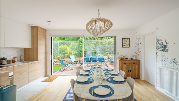 A modern kitchen with wooden cabinets, a dining area with a table set for six, and a view of a garden through glass doors. - Ti Armor - Maison à 200m de la plage (Benodet)
