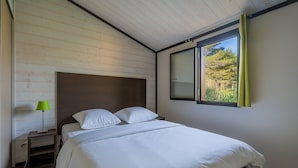 A bedroom with a wooden ceiling, a bed with white bedding, a nightstand with a lamp, and a window with a view of greenery.