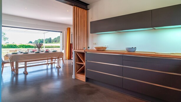 A modern kitchen with a long wooden table set for dining, a dark-colored cabinet with drawers, and a view of greenery outside. - Ti Ar Mor Braz- Maison avec grand jardin (Saint-Philibert)