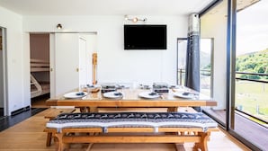 Comedor con una mesa de madera dispuesta para cuatro personas, un banco con cojines estampados, un televisor montado y vistas a un paisaje verde a través de grandes ventanales.