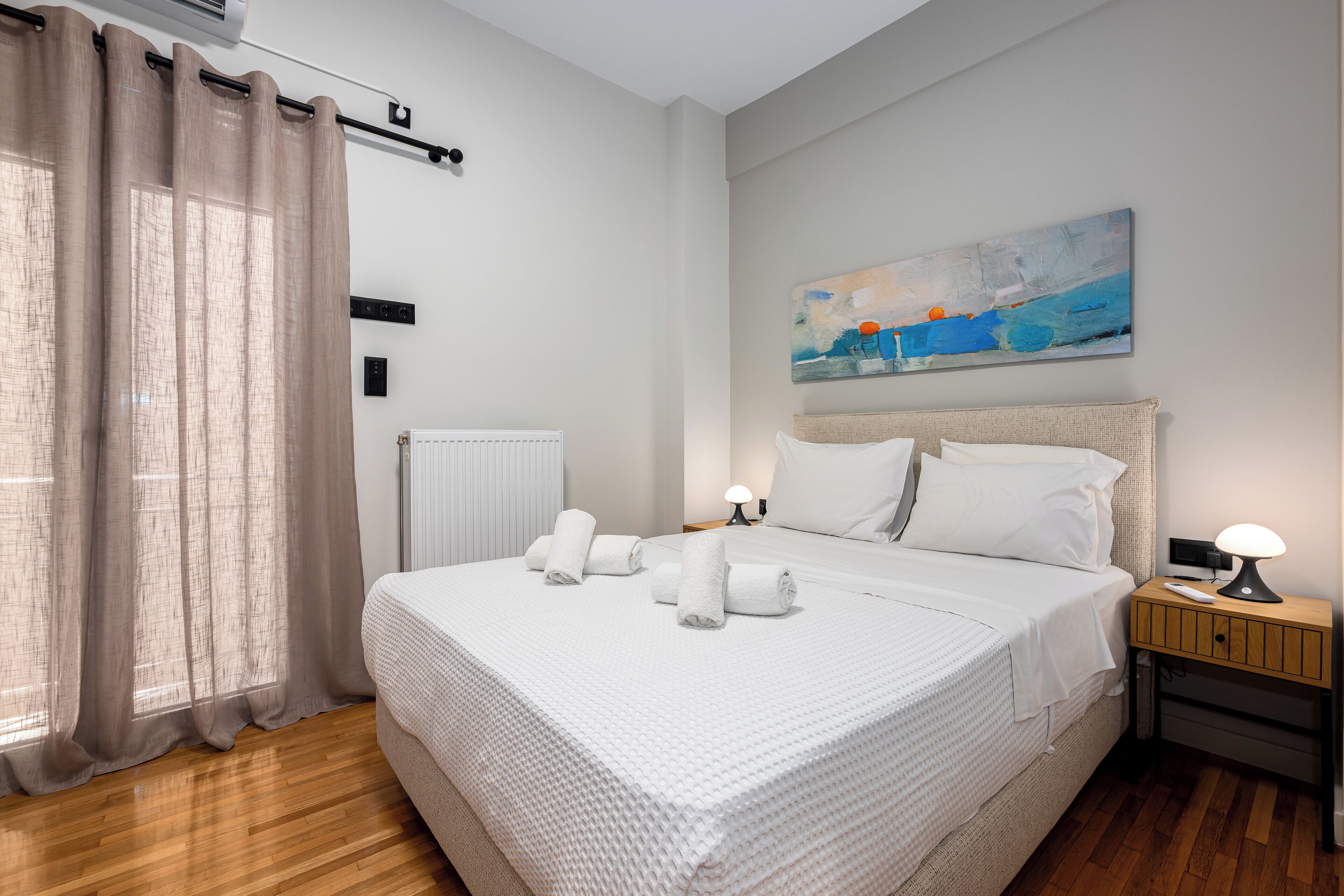 A neatly made bed with white linens and towels, a bedside table with a lamp, a painting on the wall, and a window with curtains.