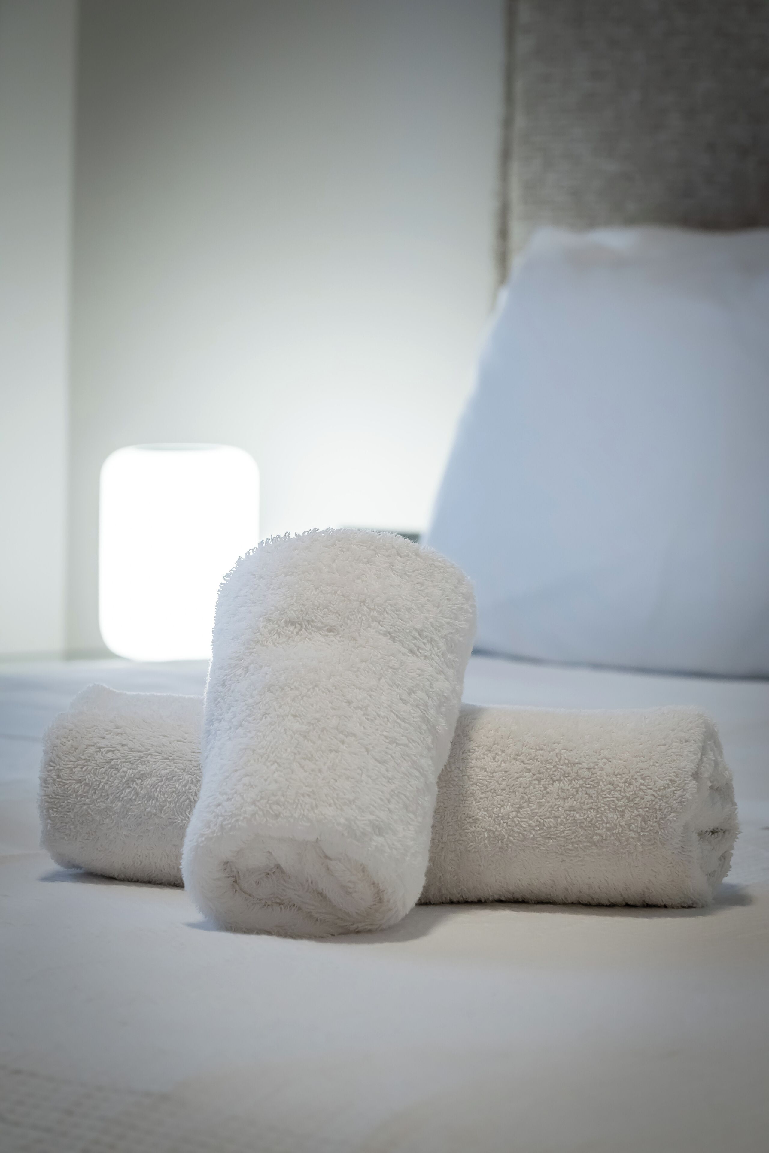 Three rolled white towels on a bed with a white headboard and a window in the background.