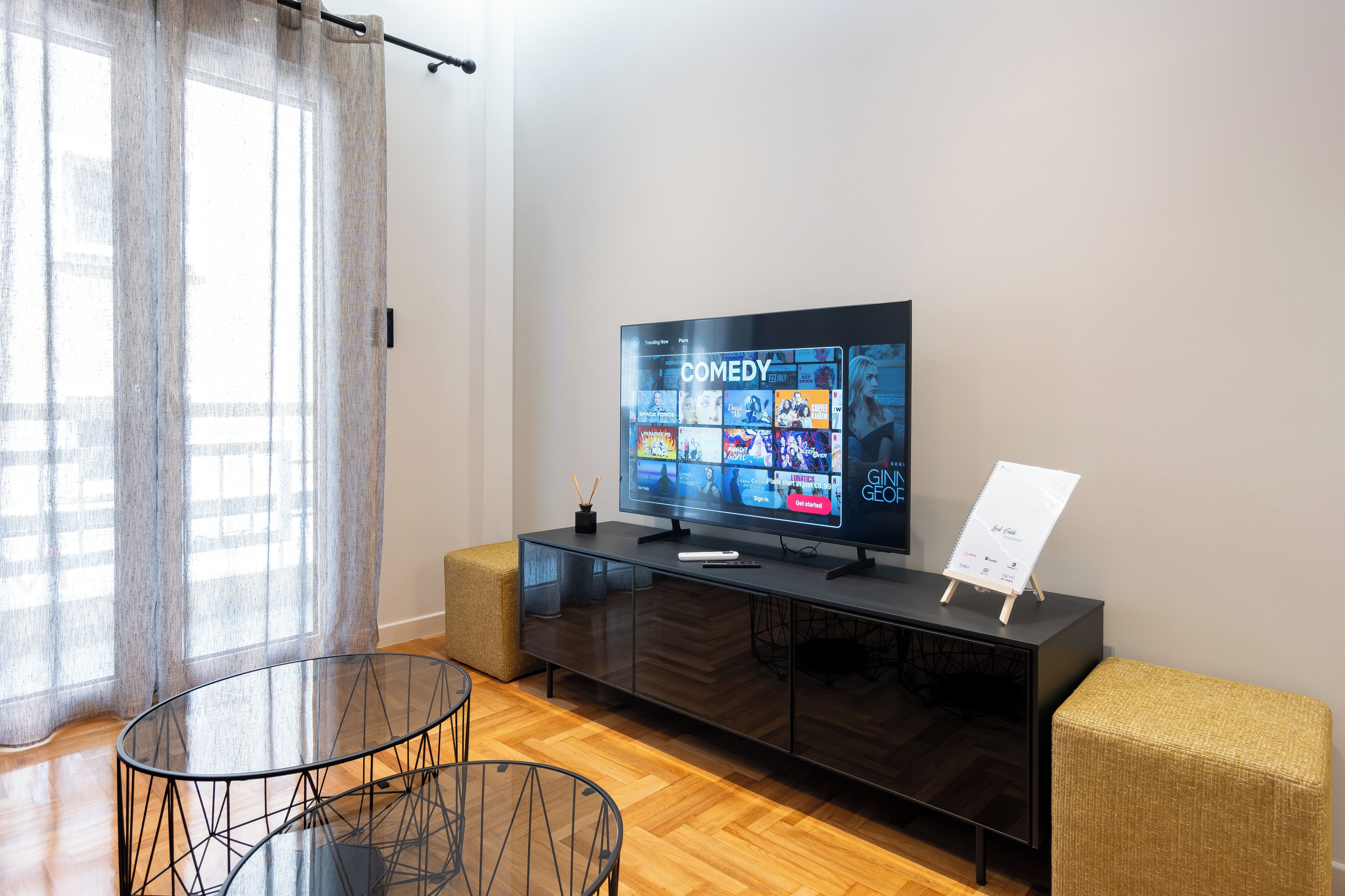 A modern living room with a flat-screen TV displaying a comedy channel, a glass coffee table, and a dark wooden cabinet with a small easel.