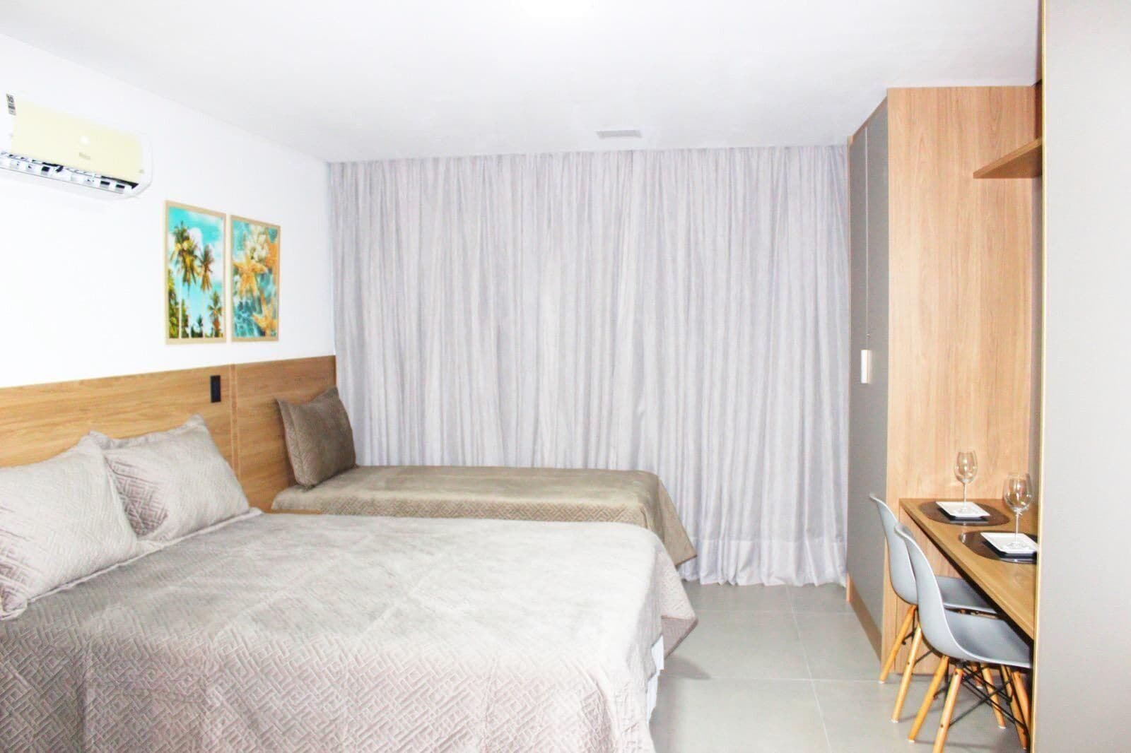 A hotel room with two beds, a wooden headboard, a dining area with a table and chairs, and a large window with sheer curtains.