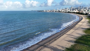 Uma praia costeira com areal, área gramada e um horizonte urbano ao fundo.