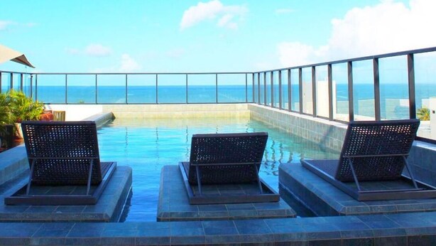 Piscina infinita no terraço, com duas cadeiras de vime e um espaço bar, com vista para o oceano.